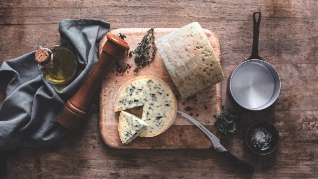 Fromage Fourme d’Ambert tranché sur une planche avec huile d’olive, poivre, thym, sel et ustensiles de cuisine sur table en bois.