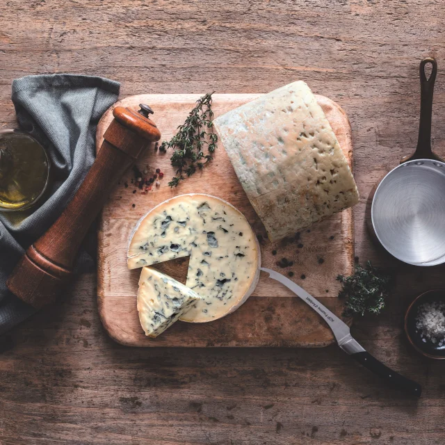 Fromage Fourme d’Ambert tranché sur une planche avec huile d’olive, poivre, thym, sel et ustensiles de cuisine sur table en bois.