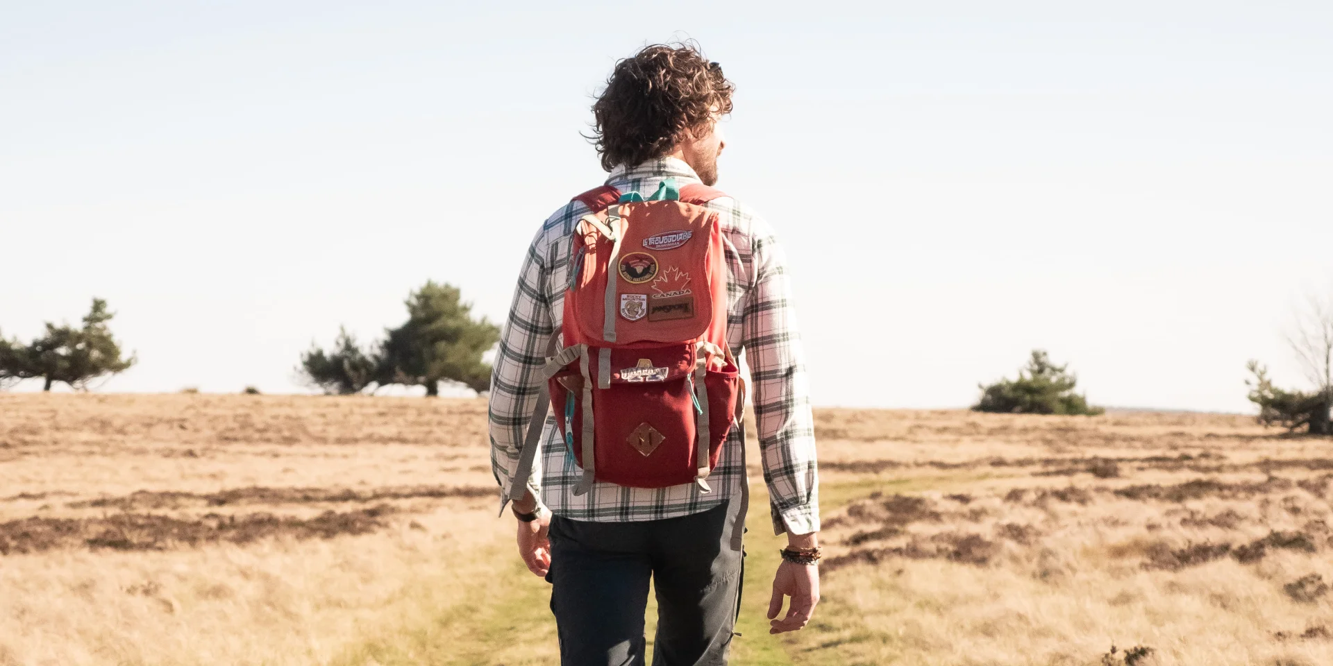 Un homme de dos avec un sac à dos rouge marche sur un sentier herbeux, entouré de champs secs et d'arbres clairsemés, sous un ciel bleu dégagé.