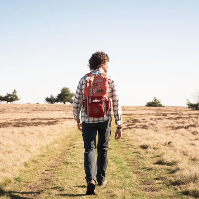 Un homme de dos avec un sac à dos rouge marche sur un sentier herbeux, entouré de champs secs et d'arbres clairsemés, sous un ciel bleu dégagé.