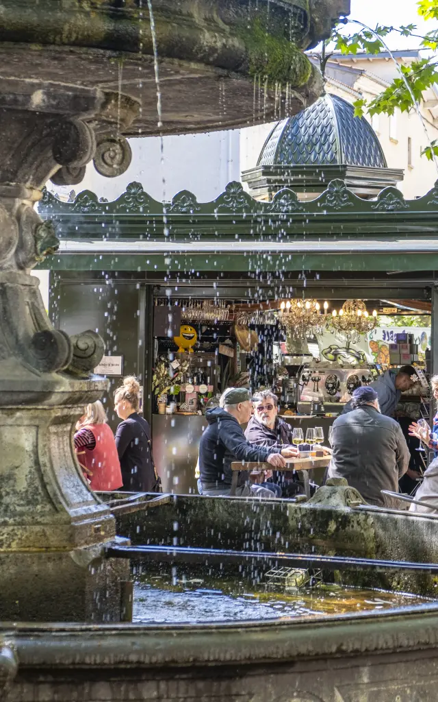 Fontaine au premier plan et kiosque animé à Billom avec terrasse bondée lors d’un après-midi d’été.