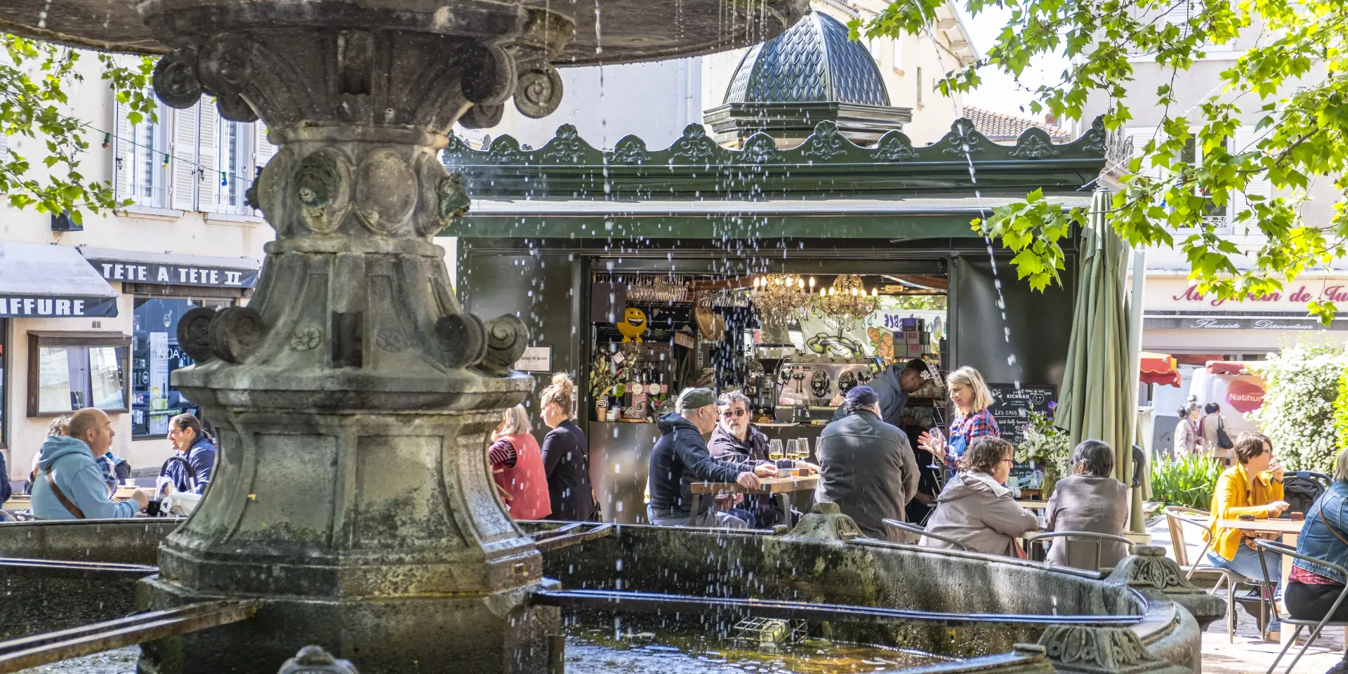 Fontaine au premier plan et kiosque animé à Billom avec terrasse bondée lors d’un après-midi d’été.